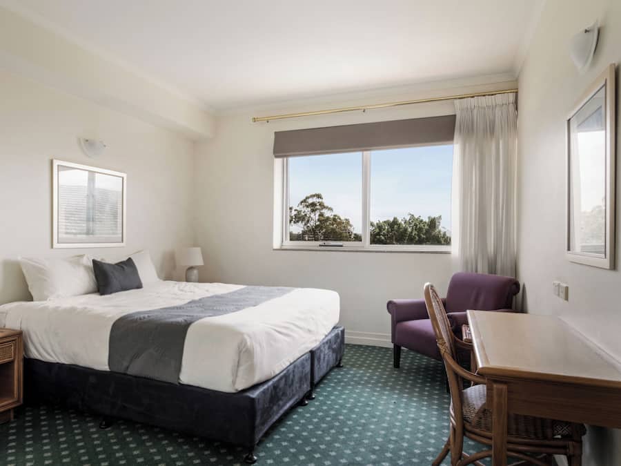 A hotel room with a large bed, a chair, a small table, and a window with a view of trees.