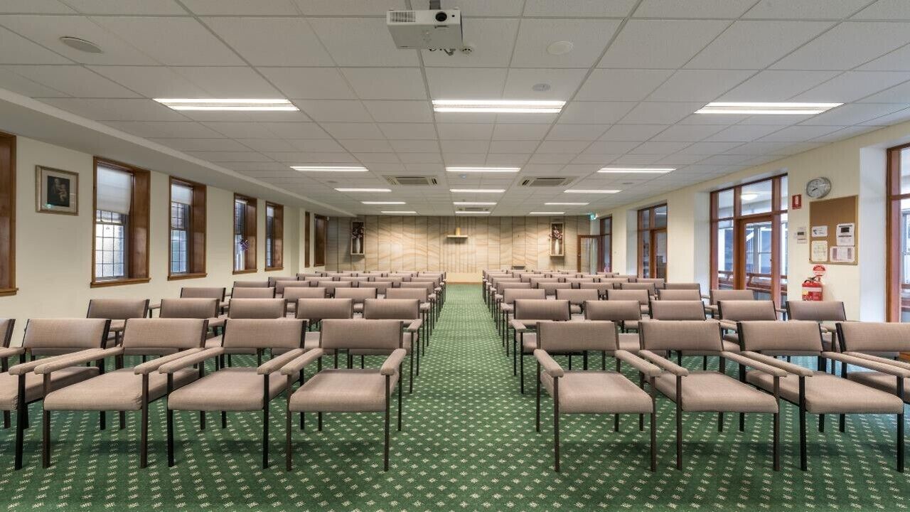 Sala de conferências com fileiras de cadeiras, um projetor e piso coberto de carpete.