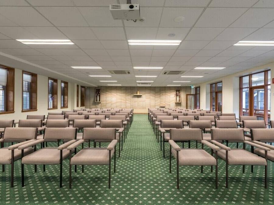 Sala de conferências com fileiras de cadeiras, um projetor e piso coberto de carpete.