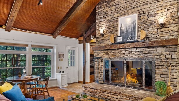 A cozy living room with a stone fireplace, wooden ceiling beams, and a dining area with a table and chairs.