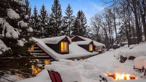 A cabin with a snow-covered roof, a fire pit, and a chair in the foreground.