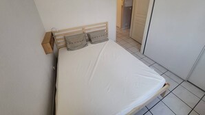 A single bed with a wooden headboard and two pillows, a white wall, and a tiled floor. - Capbreton – Clos Paisible residence in town centre with parking. (CAPBRETON)