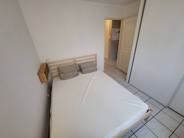 A single bed with a wooden headboard and two pillows, a white wall, and a tiled floor.
