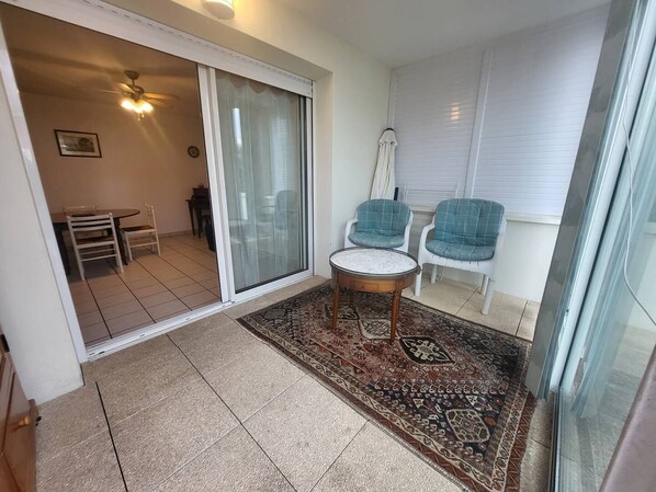 A balcony with a sliding glass door, a small round table, two chairs, a patterned rug, and a ceiling fan.