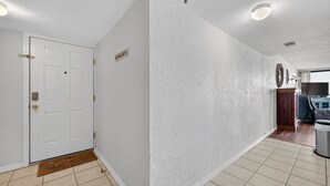 Un pasillo con una puerta blanca, un felpudo, una lámpara de pared y un timbre.
