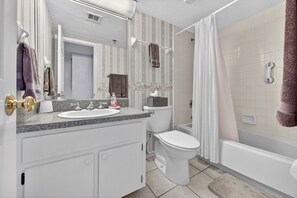 A bathroom with a white sink, toilet, and bathtub. There is a shower curtain and a towel rack with towels. - Top floor beachfront condo in peaceful NSB (New Smyrna Beach)