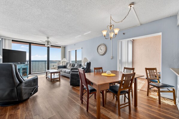 A spacious living area with a wooden dining table, comfortable seating, a ceiling fan, and a large window offering a view of the sky. - Top floor beachfront condo in peaceful NSB (New Smyrna Beach)