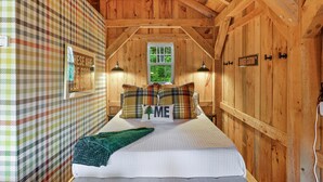 A cozy bedroom with a plaid-patterned wall, a bed with plaid pillows, a green throw blanket, and a window with a view of greenery. - Fall Adventures • Private Hot Tub • Sunday River (Bethel)