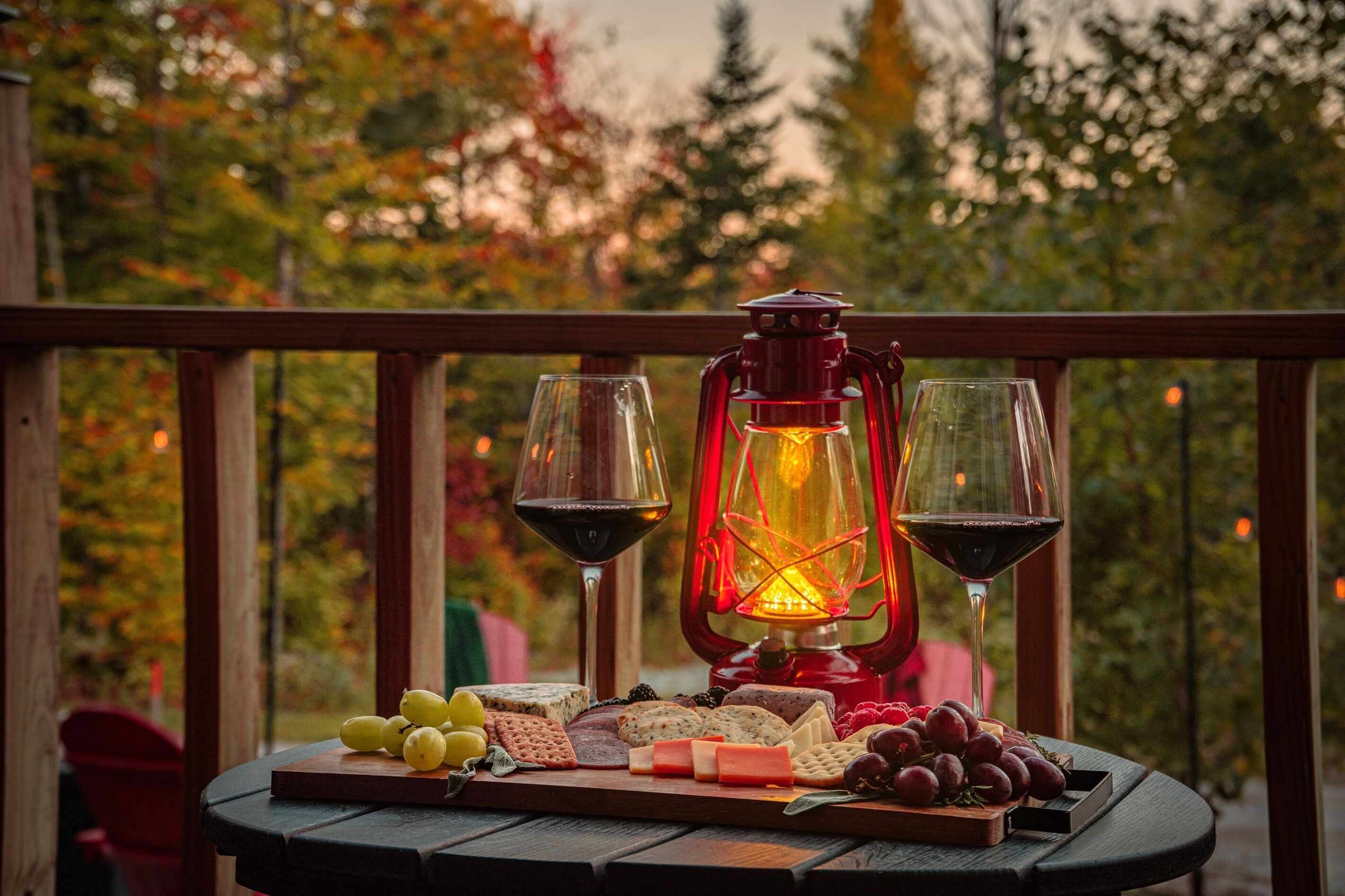 Una mesa de madera con una tabla de quesos, uvas y un farolillo rojo en un balcón con vistas a árboles.
