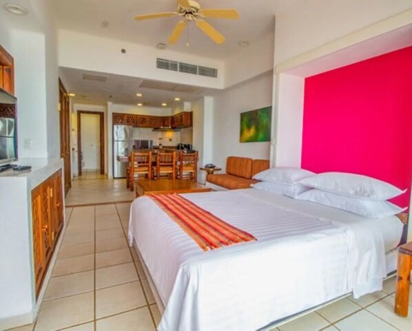 A hotel room with a large bed, a dining area with a table and chairs, and a kitchen in the background.