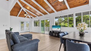 A spacious room with a high ceiling, wooden beams, a large TV, and a dining table with chairs. - Renovated 3BR w/ AC, Vaulted Ceilings & Lanai Ocean View Near Airport (Kailua-Kona)