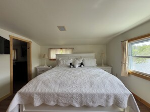 Quarto com uma cama grande, duas mesas de cabeceira, uma janela com cortinas e uma porta que dá para outro cômodo.