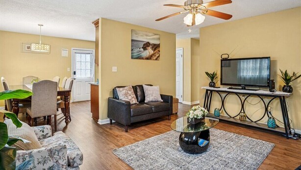 A living room with a sofa, a coffee table, a TV on a stand, a ceiling fan, and a dining area with chairs and a table. - Spacious Family Stay, Large Dining, 3 Cozy Bedrooms, Home Near Buckroe Beach (Hampton)