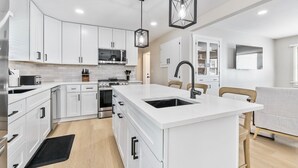 A modern kitchen with white cabinetry, a central island with a sink, a microwave, and a stove. There is a dining area with a gray sofa and a TV mounted on the wall. - Fox Island Lakefront Retreat (Lakeview)