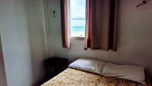 A small, simple room with a bed, a window with curtains, and a small table. - Neri House - Ocean View, Beach Front (Cidade Ocian Praia Grande)