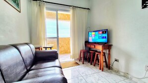 A living room with a leather sofa, a TV on a wooden stand, and a sliding glass door leading to an outdoor area. - Neri House - Ocean View, Beach Front (Cidade Ocian Praia Grande)