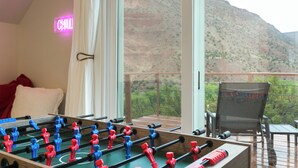 Una habitación con mesa de fútbol, una silla, ventana con vistas a montañas y un letrero de neón que dice 'CHILL'.