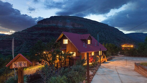 Una casa de dos pisos con techo rojo, rodeada por un jardín ajardinado y con montañas al fondo.