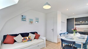 Ein modernes Wohnzimmer mit einem weißen Sofa, bunten Kissen, einem Esstisch mit Tellern und Gläsern sowie einer Küchenzeile mit dunklen Schränken.