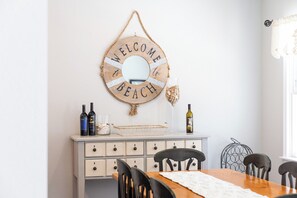 A dining area with a wooden table, chairs, a white cabinet with drawers, a decorative welcome sign, and bottles on the cabinet. - Coast to Coast:Dog Friendly, Private Pool, Hot Tub, Community Amenities (Corolla)