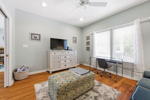 A room with a TV on a dresser, a desk with a chair, a patterned ottoman, and a window with blinds. - Coast to Coast:Dog Friendly, Private Pool, Hot Tub, Community Amenities (Corolla)