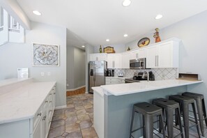 A modern kitchen with white cabinets, a central island with stools, a refrigerator, a microwave, and a coffee maker. - Coast to Coast:Dog Friendly, Private Pool, Hot Tub, Community Amenities (Corolla)