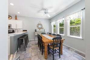 A kitchen with white cabinets, a dining table with chairs, a ceiling fan, and a view of greenery through large windows. - Coast to Coast:Dog Friendly, Private Pool, Hot Tub, Community Amenities (Corolla)