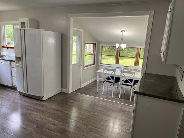 A kitchen with a refrigerator, a dining area with a table and chairs, and a view of the outdoors through large windows.