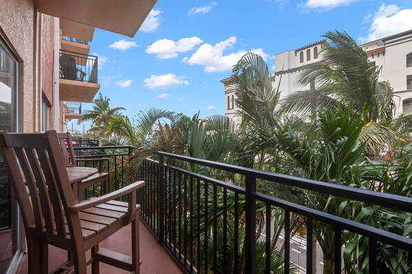 A balcony with a wooden chair, a railing, and a view of palm trees and buildings.