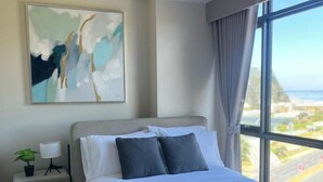 Una camera d'albergo moderna con un letto grande, un comodino con una lampada e una pianta, un quadro al muro e vista sulla città attraverso un'ampia finestra.