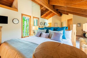 A bedroom with a large bed, wooden beams, a clock, and a window with a view of trees.