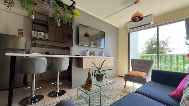 A modern living room with a sofa, a glass coffee table, and a bar area with stools. There is a large window providing a view of the outdoors. - 10 minutes from International Airport, Vespucio business sector. (Renca)