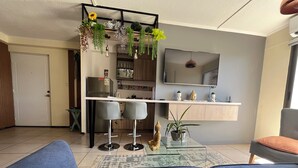 A modern living room with a sofa, a glass coffee table, and a kitchen area with a TV and plants. - 10 minutes from International Airport, Vespucio business sector. (Renca)