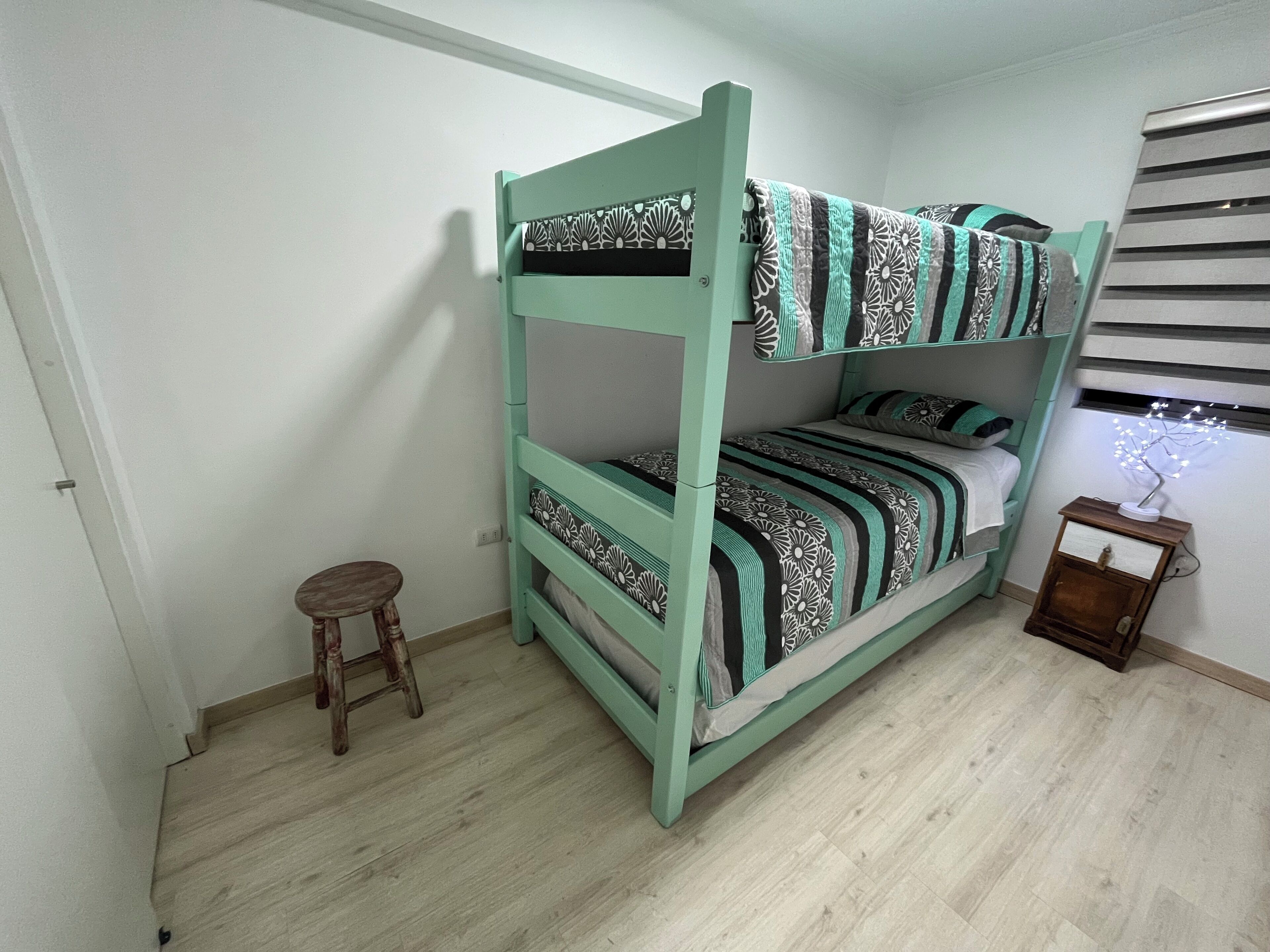 A bunk bed with a patterned bedspread, a small wooden stool, and a nightstand with a decorative item.