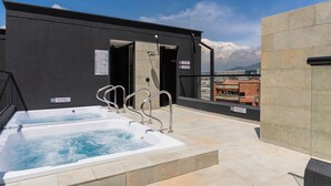 Een terras op het dak met twee whirlpools, een zwarte muur en een uitzicht op gebouwen op de achtergrond.