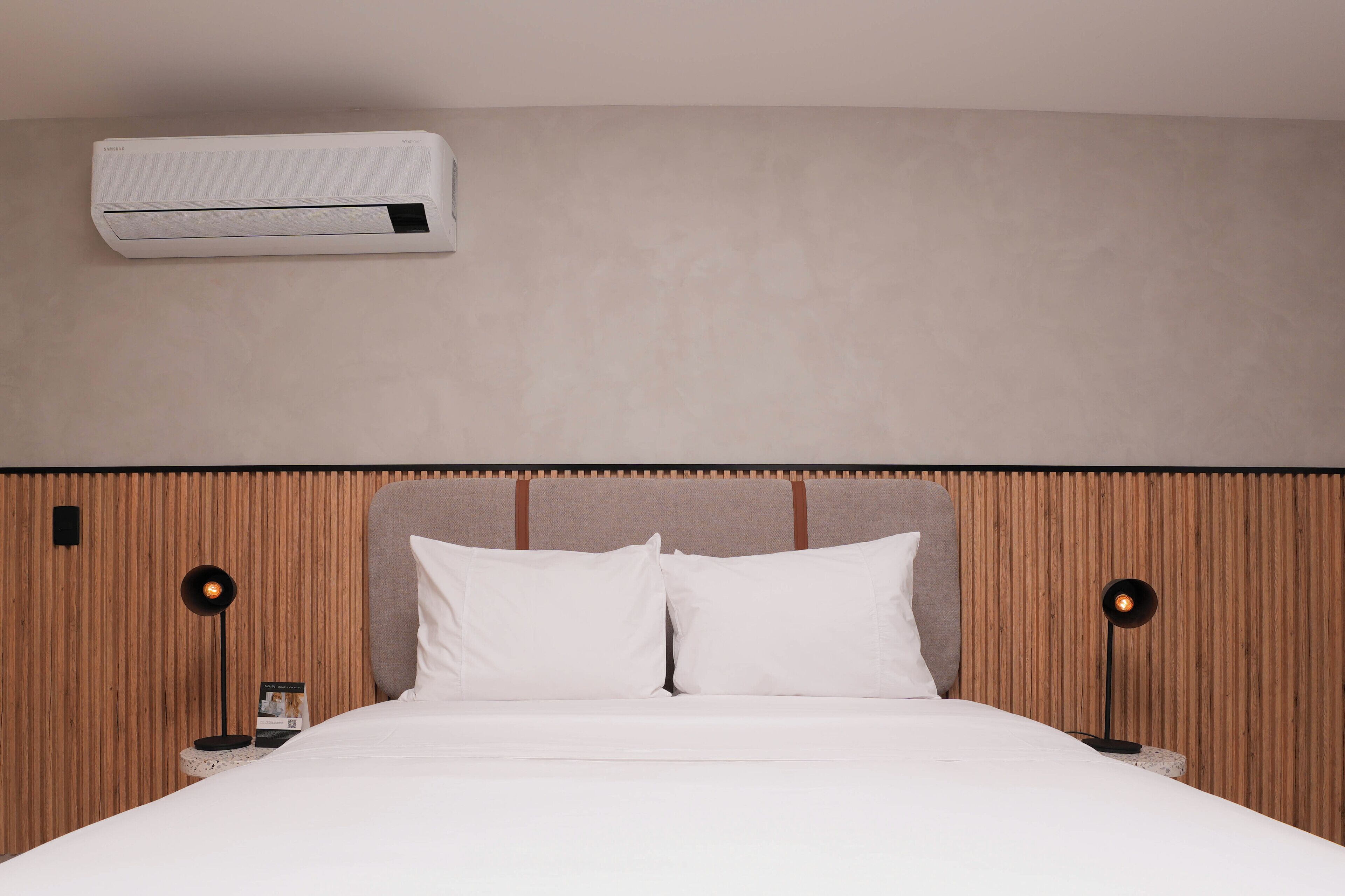 Een moderne hotelkamer met een houten hoofdbord, witte beddengoed en een wandairconditioner.