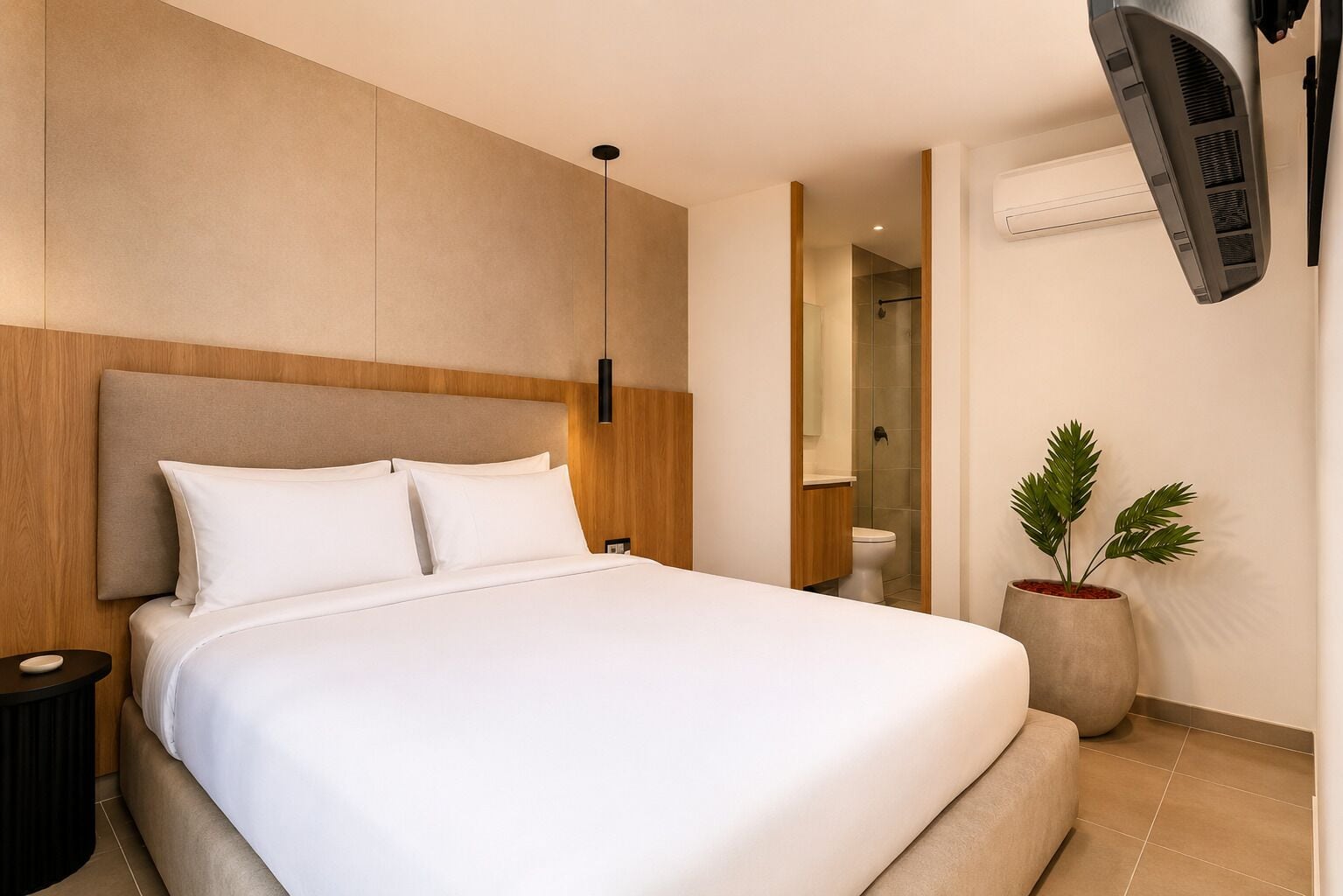 Habitación de hotel con una cama grande, una mesita de noche, una planta en maceta y un baño visible a través de una puerta abierta.