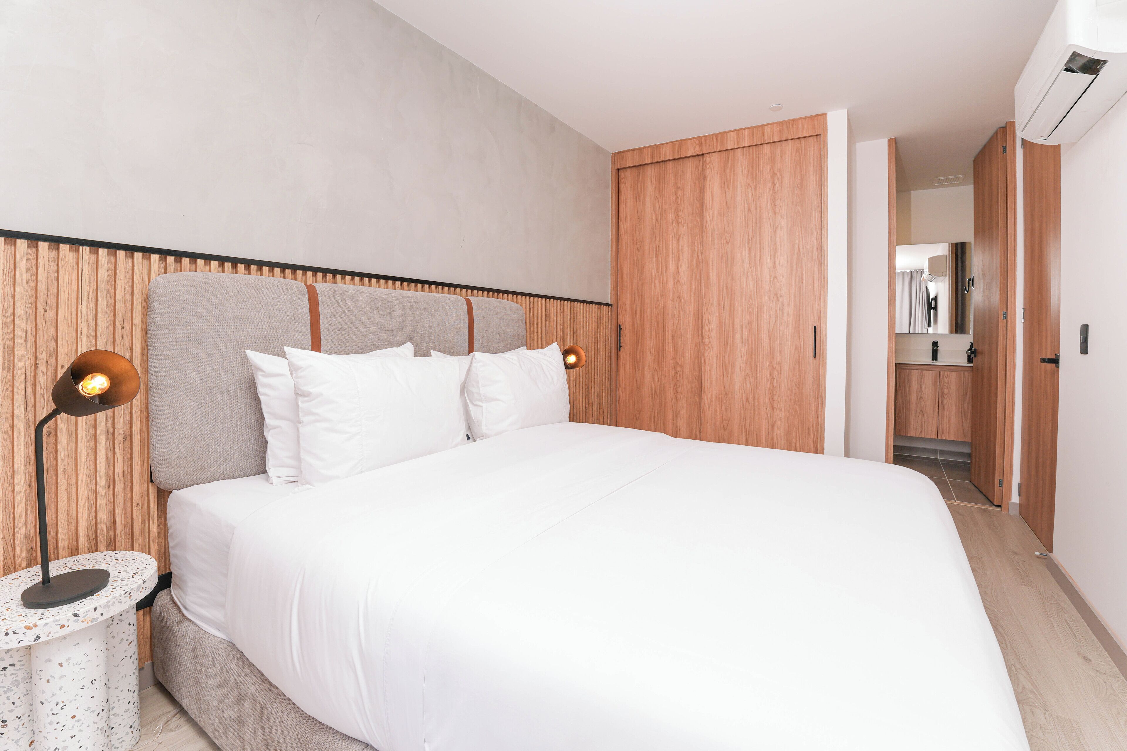 A modern bedroom with a large bed, wooden headboard, and a round nightstand with a lamp.