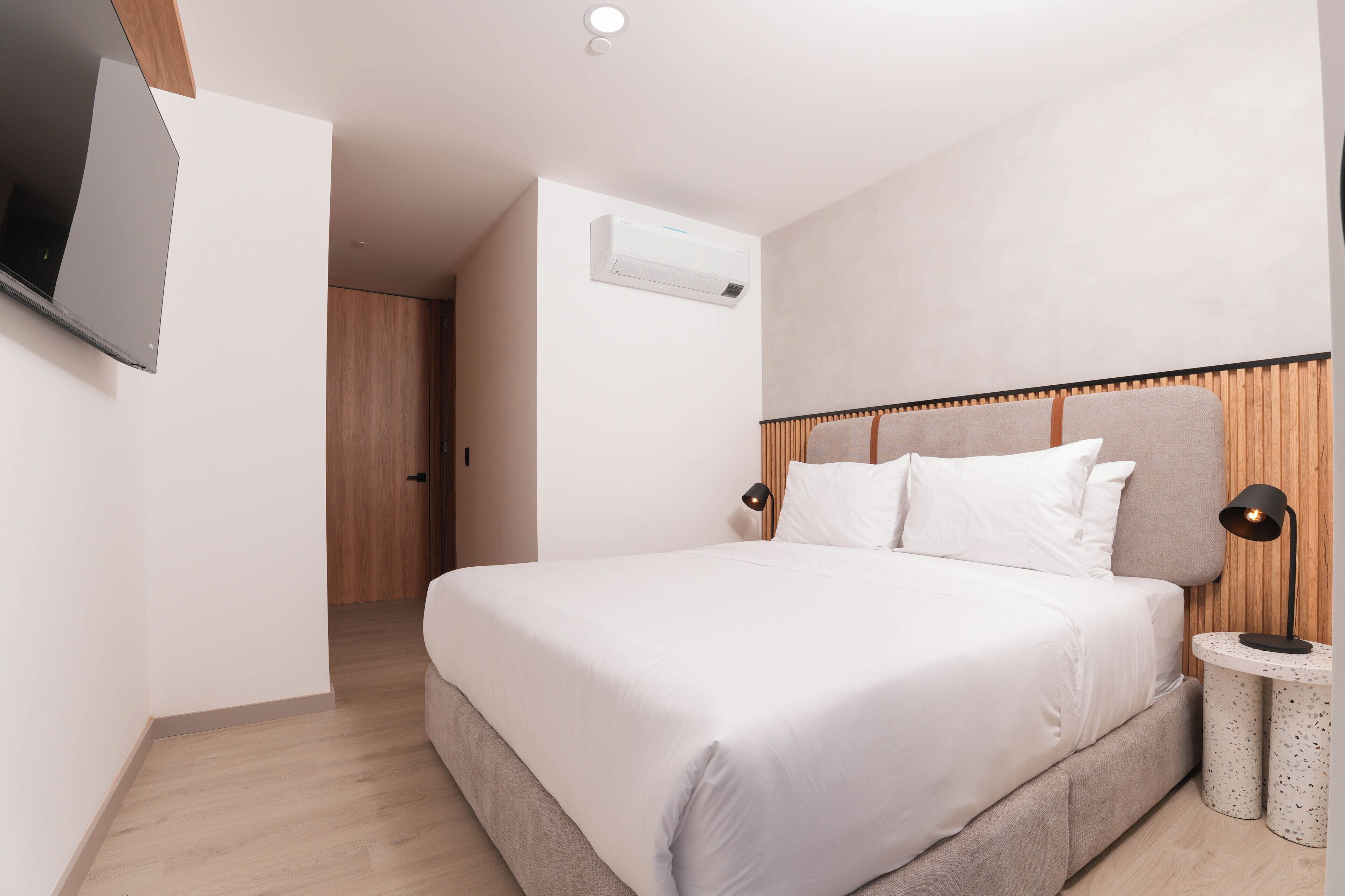 A modern hotel room with a large bed, a flat-screen TV mounted on the wall, and a small side table with a lamp.