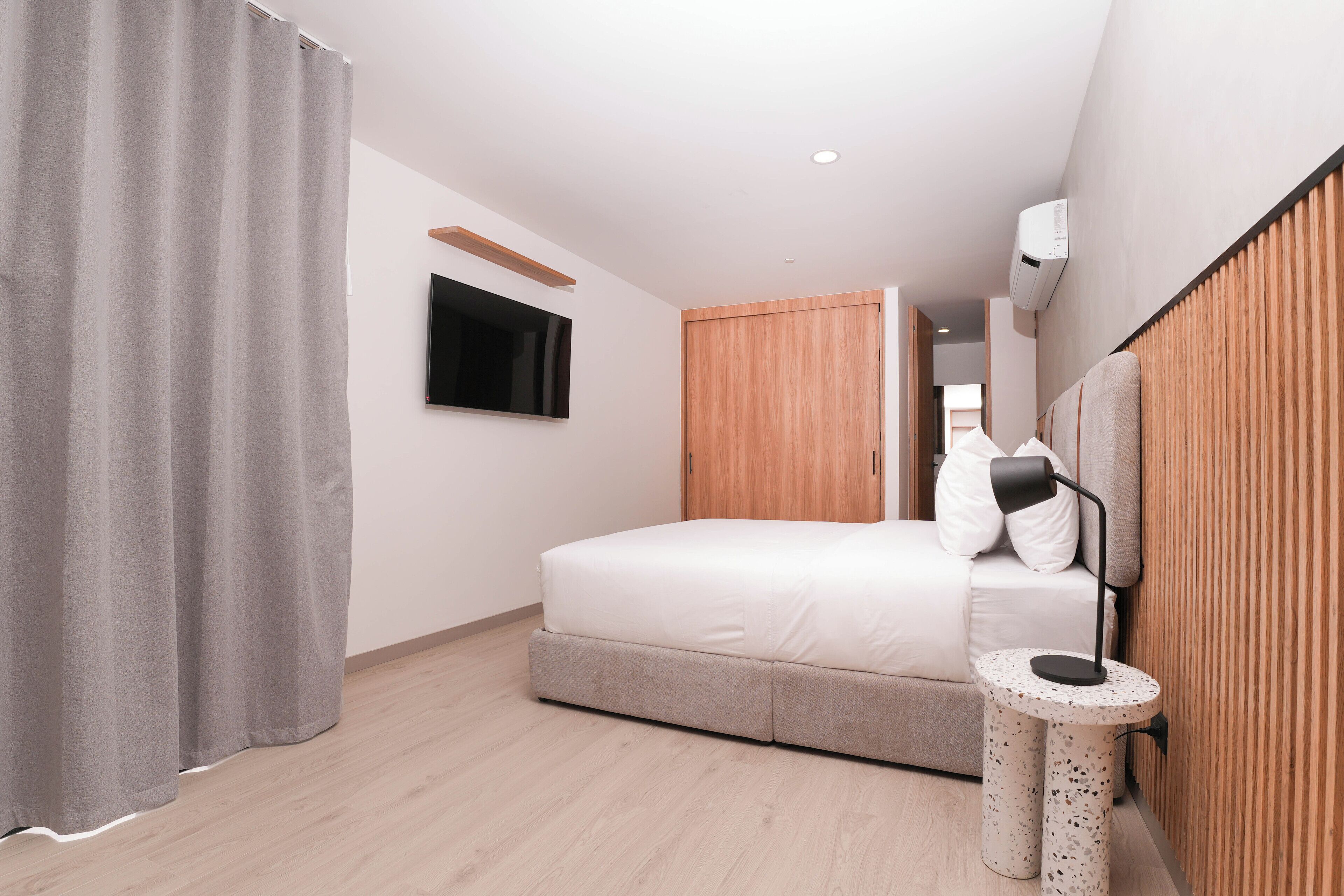A modern hotel room with a bed, a TV mounted on the wall, a wooden wardrobe, and a round side table with a lamp.