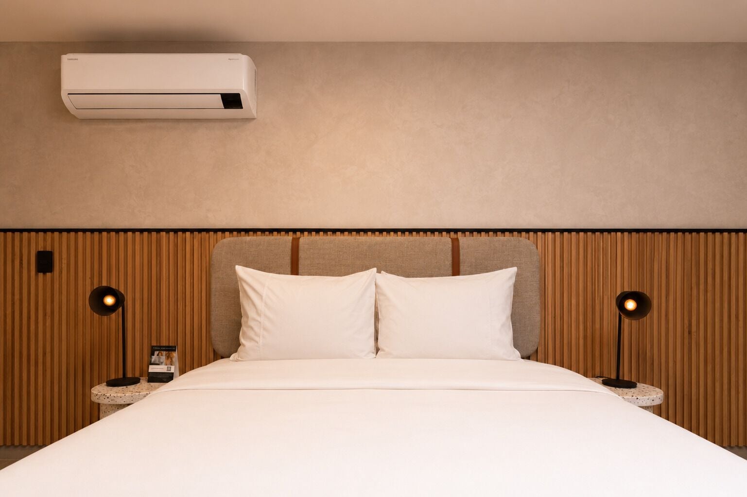 A hotel room with a bed, two bedside lamps, an air conditioner, and a wooden panel wall.