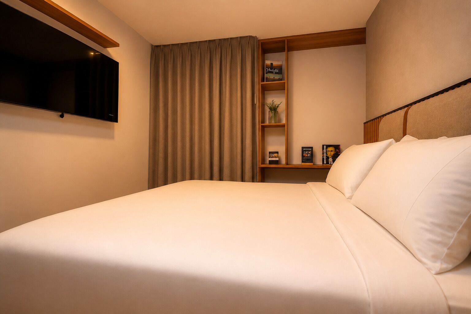 A hotel room with a large bed, a flat-screen TV mounted on the wall, a bookshelf with books and decorative items, and a window with curtains.