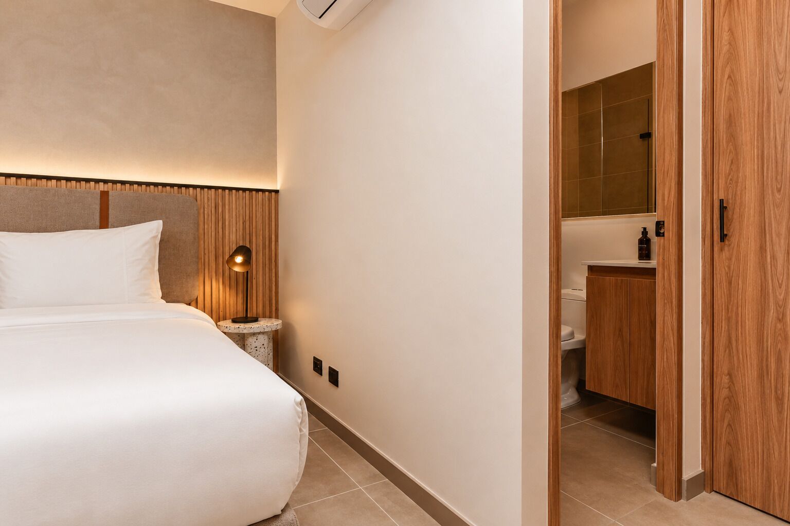 A hotel room with a bed, bedside table, lamp, and a view into the bathroom.
