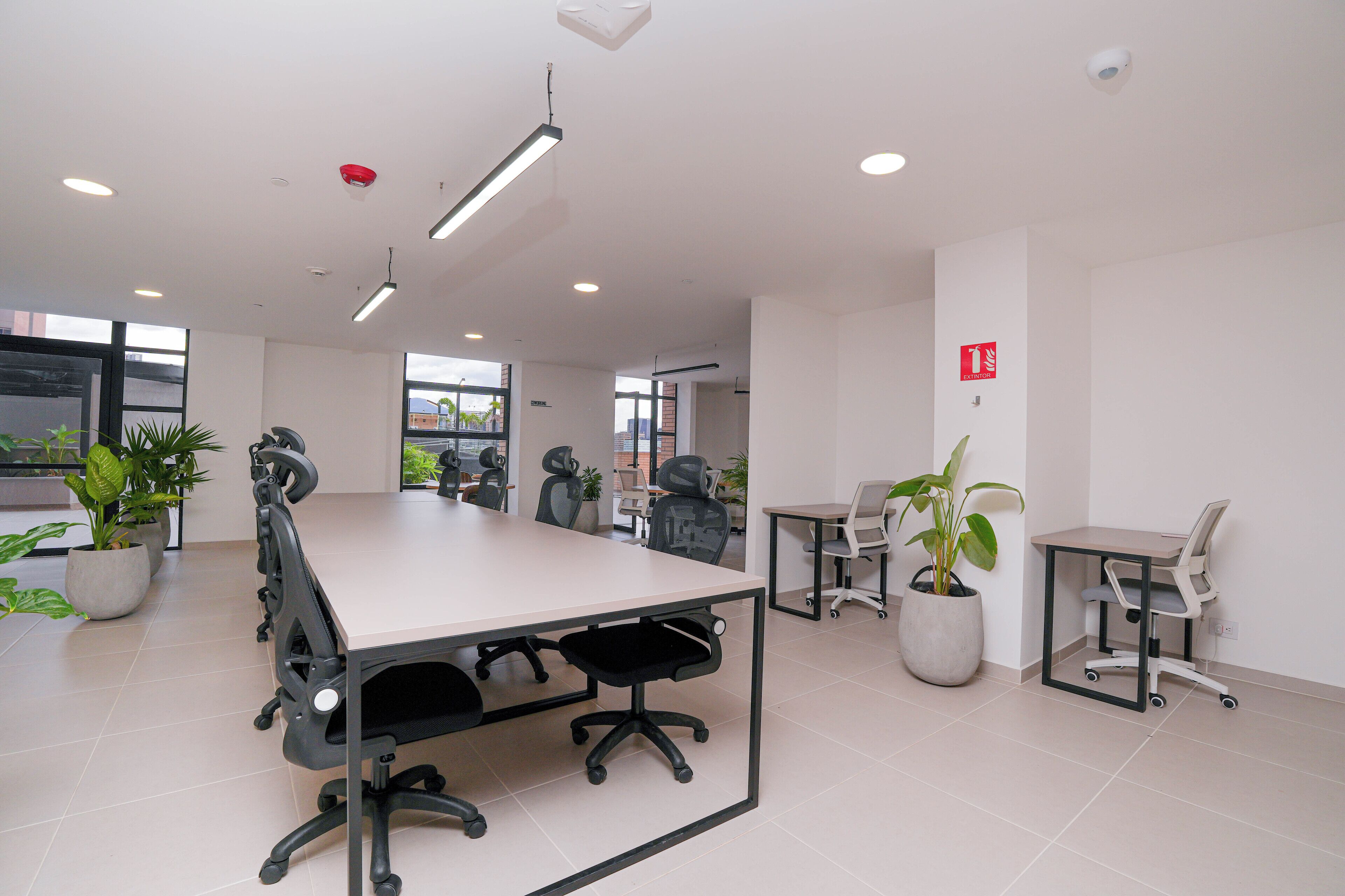 A modern office space with a large meeting table, several office chairs, potted plants, and a fire alarm on the wall.
