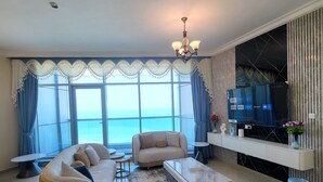 A modern living room with a sofa set, a coffee table, and a TV. The room has large windows with curtains and a view of the sea. - Sunrise and sunset sea views luxury 2Bedrooms apartments. (Ajman)