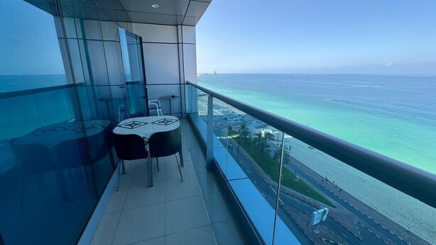 A balcony with a table and chairs, offering a view of the ocean and a road with buildings. - Sunrise and sunset sea views luxury 2Bedrooms apartments. (Ajman)