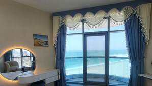 A hotel room with a bed, a desk, a chair, a window with a view of the beach, and a balcony with curtains. - Sunrise and sunset sea views luxury 2Bedrooms apartments. (Ajman)