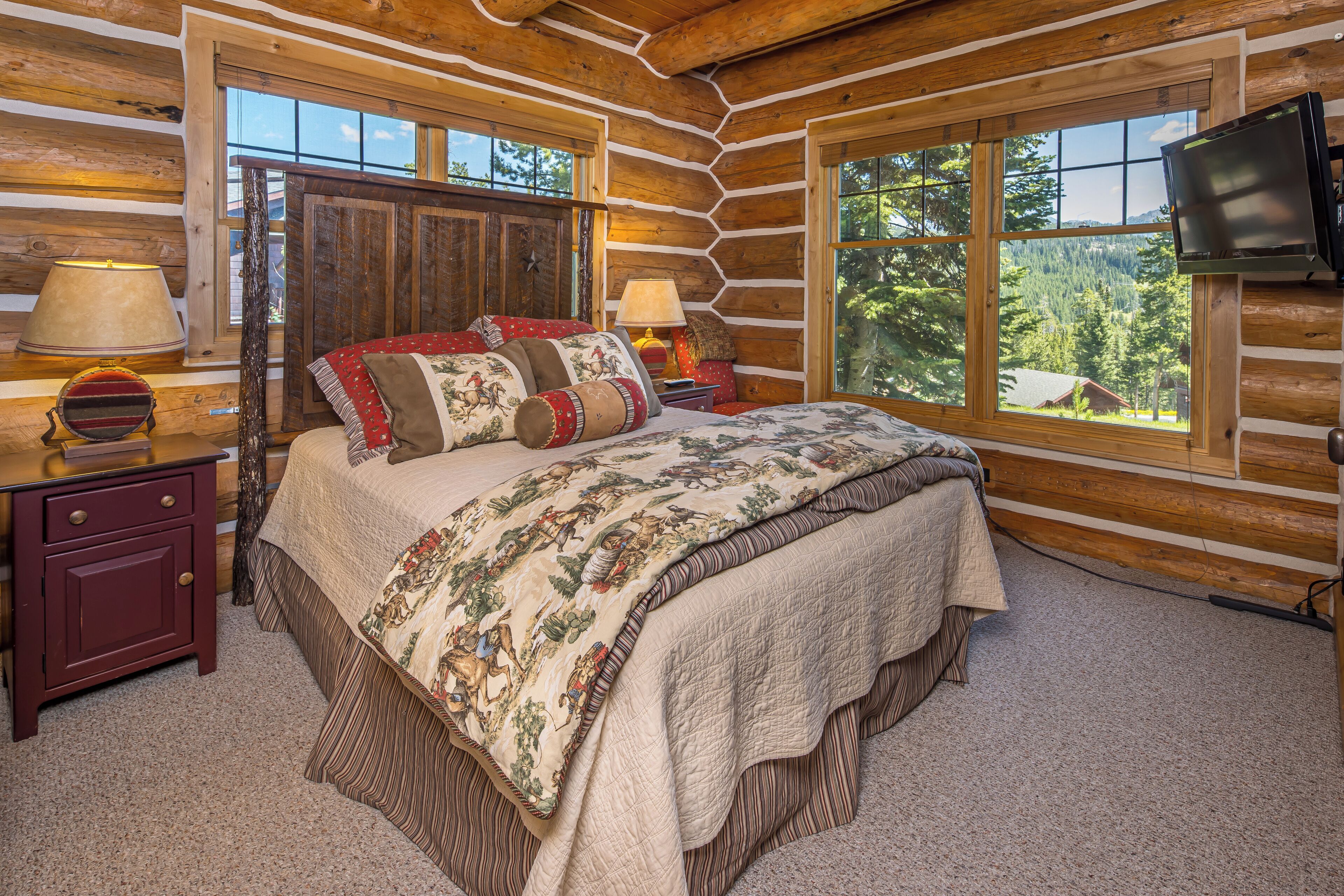 A log cabin bedroom with a large bed, two bedside tables, a TV, and a window with a forest view.