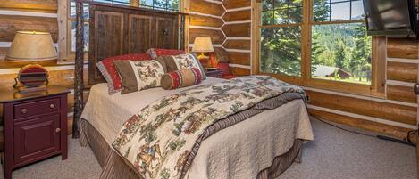 A log cabin bedroom with a large bed, two bedside tables, a TV, and a window with a forest view.