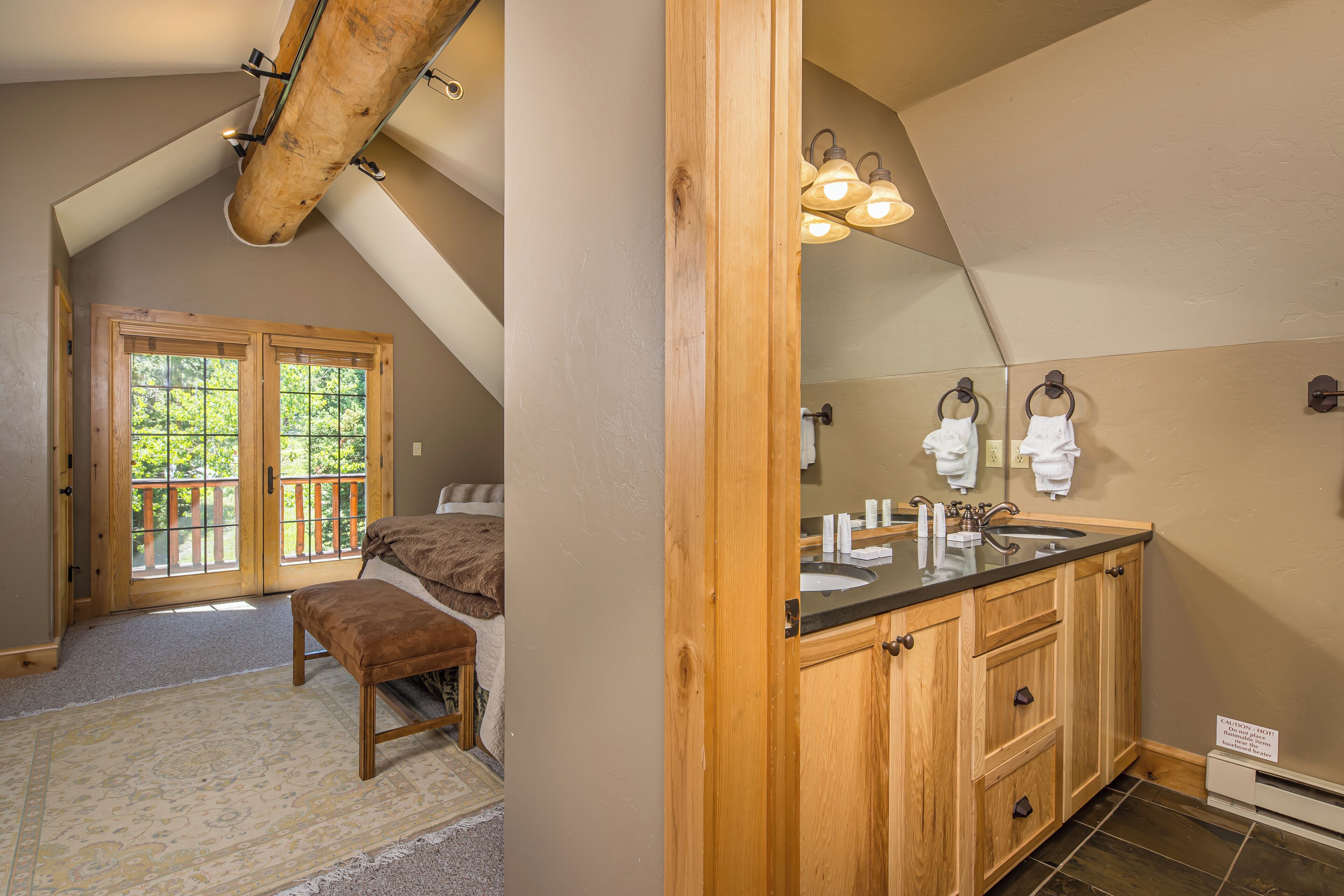 A bedroom with a large window, a wooden bench, and a bathroom with a sink and mirror.